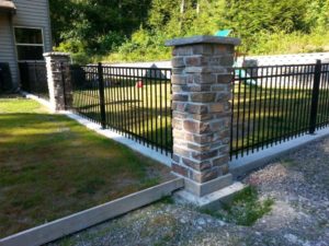 A backyard with a black metal fence and stone pillars encloses a grassy area with trees in the background. A blue and green playground set is visible inside the yard. The fence is bordered by a gravel path on the outside.