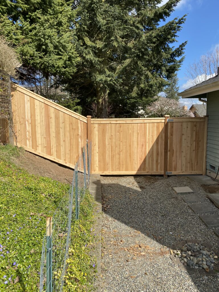 A new wooden fence spans across a sloped, gravel pathway. Sunlight casts shadows on the fence and the surrounding area. Trees and grass-lined areas are visible in the background under a clear blue sky.