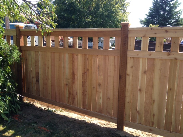 A wooden privacy fence with vertical planks and a decorative top row of square openings is shown. It stands in a grassy area with trees and bushes nearby, under a sunny sky.