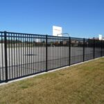 A black metal fence surrounds an outdoor basketball court with two visible hoops. The court is located on a grassy area under a clear blue sky.