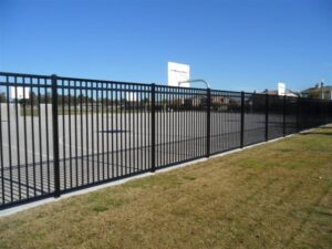 A black metal fence surrounds an outdoor basketball court with two visible hoops. The court is located on a grassy area under a clear blue sky.